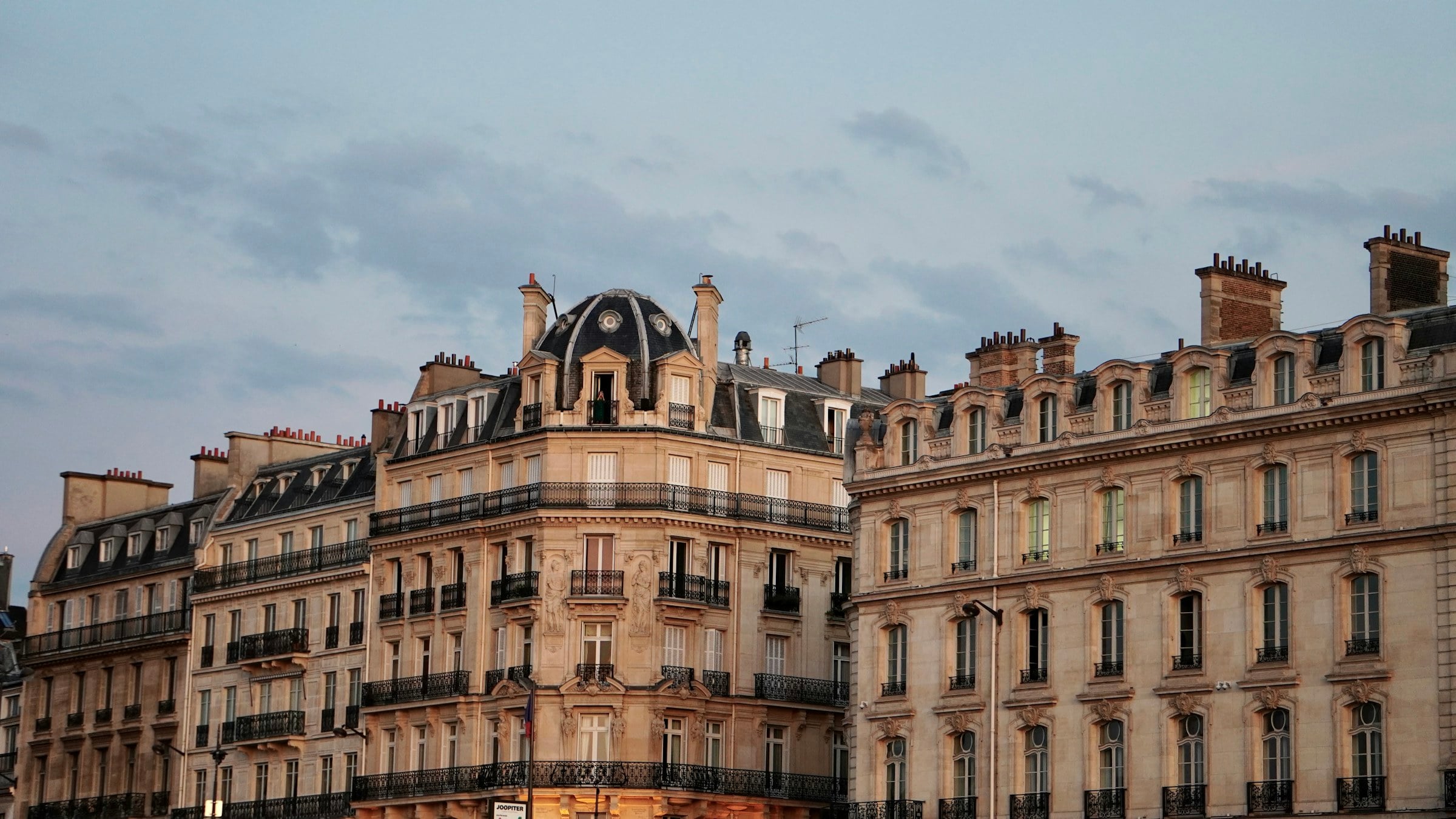 Immeuble haussmannien d'angle au crépuscule : façade en pierre de taille, dôme en ardoise coiffé de lucarnes et balcons filants en fer forgé.