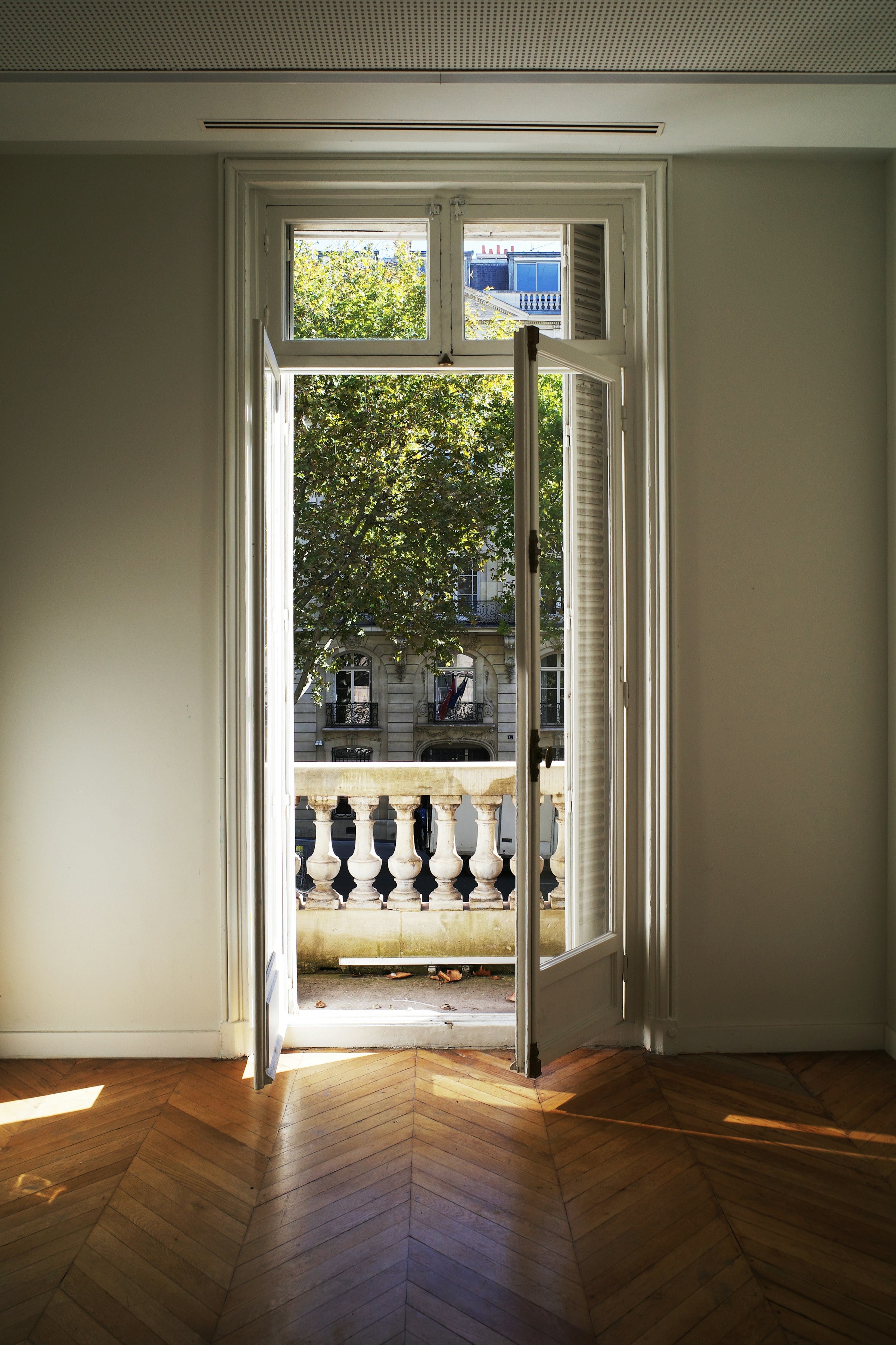 Séjour d'appartement parisien au parquet en point de Hongrie, porte-fenêtre ouverte sur un balcon et la façade d'en face.